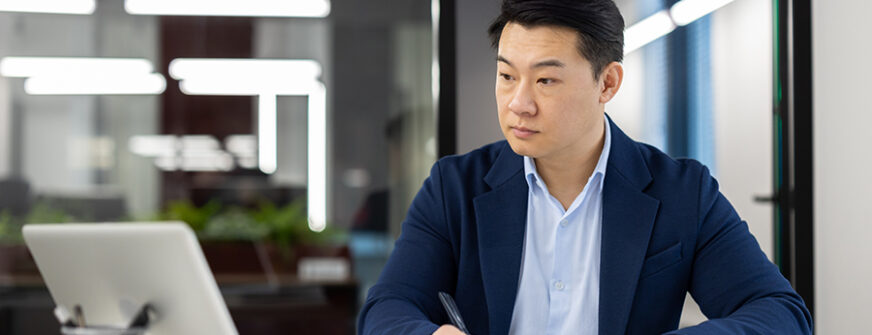 Professional Asian businessman concentrating on work, reviewing documents with laptop in a well-lit modern office.