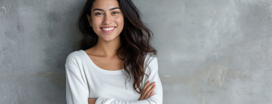 Portrait of a young latin woman with pleasant smile and crossed arms isolated on grey wall with copy space.